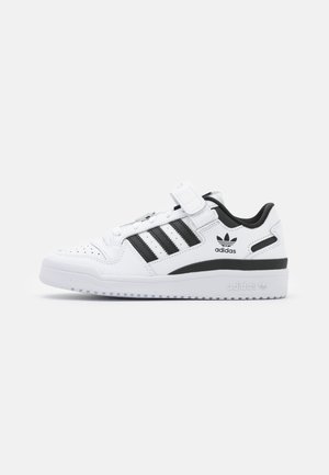 Sneaker bianca Adidas con tre strisce nere, colletto del tallone nero, cinturino in velcro e punta traforata su sfondo bianco.