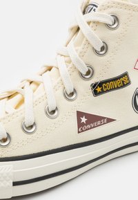 Baskets montantes en toile beige avec semelle en caoutchouc noir, œillets en argent et divers patchs colorés incluant le logo "Converse" et des motifs d'étoiles.