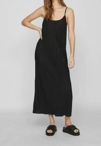 Zwarte geplooide maxi jurk met dunne banden, gemaakt van lichte stof. Model draagt zwarte slide sandalen en staat met één hand op de heup.