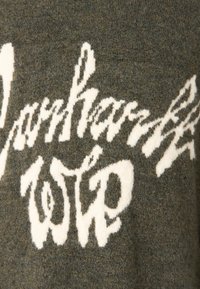 Sötétzöld kötött pulóver fehér, dőlt betűkkel. Puha tapintású, enyhén laza szövésű. A betűk szélessége és magassága változó.