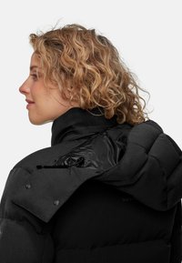 Svart isolerad puffjacka med hög krage, glansigt foder, knappdetaljer och ficka med dragkedja på axeln. Strukturerad tygfinish.