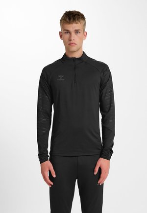 Jeune homme portant un haut de sport noir à manches longues avec fermeture éclair mi-longue et un pantalon noir assorti, debout devant un fond blanc uni.