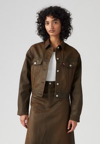 Brun dongeri jakke med sølvfargede trykknapper, to brystlommer og cropped design. Brukt over en hvit topp, kombinert med et matchende brunt skjørt.