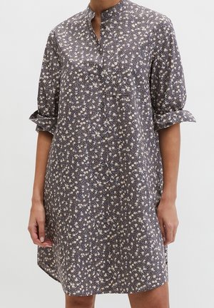 Femme portant une robe ample boutonnée avec un col montant, des manches retroussées et un petit motif floral blanc sur un tissu foncé.