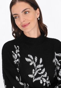 Pulover negru din tricot, cu modele de frunze din paiete argintii pe partea din față. Design cu guler înalt, textură moale și croială lejeră.