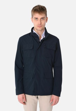 Giovane uomo che indossa una giacca blu scuro con molte tasche a pattina e con cerniera, in piedi davanti a uno sfondo chiaro e uniforme.