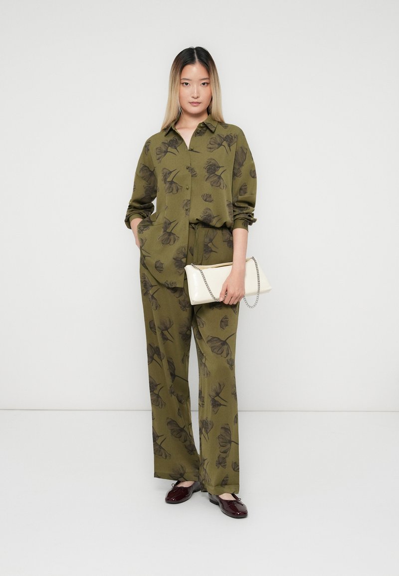 Frau in olivgrünem Blumenhemd und Hose, die eine weiße Clutch mit Kette hält, trägt dunkelrote Ballerinas und steht vor einem weißen Hintergrund.