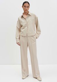 Donna con i capelli scuri raccolti in un chignon, che indossa una camicia beige con bottoni, pantaloni a gamba larga e scarpe coordinate, in piedi davanti a uno sfondo neutro.