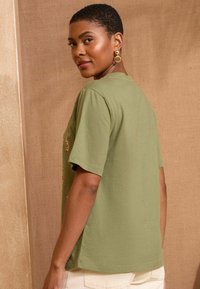 Grünes Baumwoll-T-Shirt mit kurzen Ärmeln, Rundhalsausschnitt und dezenten seitlichen Schlitzen; mit gestickten Details auf der Vorderseite.