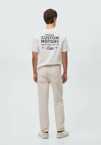 Bijela majica sa crnim natpisom "CUSTOM MOTORS PARTS AND SERVICE" na leđima, u kombinaciji sa svijetlosmešnim hlačama i svijetlim cipelama.