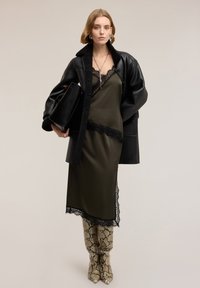 Oversize svart läderkappa över en olivgrön satinklänning med spetskant, kombinerad med knähöga ormskinnsstövlar, håller en svart väska.