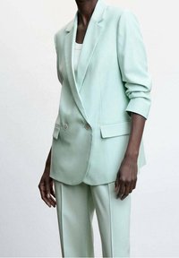 Personne portant un costume sur mesure vert menthe pâle avec un blazer croisé et un pantalon droit assorti, debout devant un fond uni.