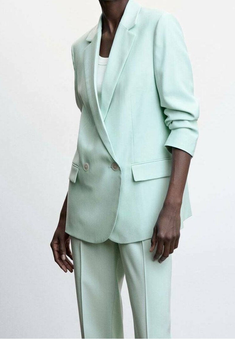 Personne portant un costume sur mesure vert menthe pâle avec un blazer croisé et un pantalon droit assorti, debout devant un fond uni.