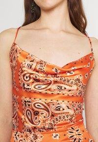Satinstopper in Orange mit einem Cowl-Ausschnitt, der mit schwarz-weißen Paisleymustern verziert ist. Dünne verstellbare Träger betonen das Design.