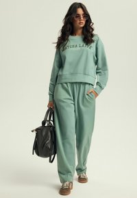 Jasnozielony, krótki sweatshirt z napisem "Matcha Latte", zestawiony z dopasowanymi zwężającymi się spodniami dresowymi. Modelka trzyma czarną torebkę, nosząc okrągłe okulary przeciwsłoneczne.