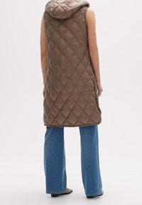 Huvtröja, ärmlös, vadderad väst i brunt med en diamantmönster och siddragkedjor, kombinerat med vida blå jeans.