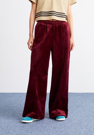 Persoon draagt wijde bordeauxkleurige fluwelen broek, een cropped beige t-shirt met zwarte strepen en teal slip-on schoenen, staand op een grijs tapijt.