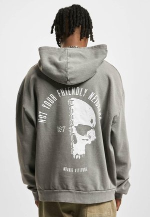Persona che indossa una felpa grigia con cappuccio abbassato, con grafica di teschio bianco e testo "Not Your Friendly Neighbor", "Thug Life" e "Meanie Attitude."