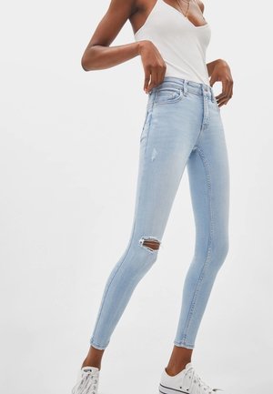 Kvinde iført lyseblå revnede skinny jeans, hvid tanktop og hvide sneakers, står med hænderne nær taljen.