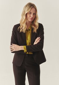 Sort tætsiddende blazer over en grøn skjorte med knapper, med matchende bukser. Fremhævet af en ring på venstre hånd og struktureret hår. Enkel baggrund.