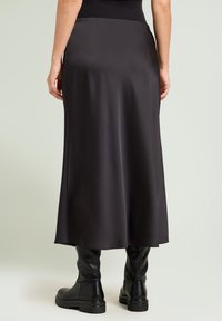 Schwarzer Satin-Maxi-Rock mit glatter Textur, der einen Taillengummi und einen subtilen Fall aufweist. Kombiniert mit eleganten schwarzen knielangen Stiefeln.