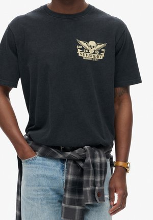 Sort bomulds-T-shirt med grafik foran, der viser en kranie og vinger i guld, kombineret med lyseblå denimjeans og en grå ternet skjorte bundet om livet.