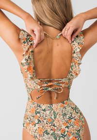 Vrouw die goudkleurige ketting verstelt, draagt een bloemenbadpak met gerimpelde schouderbandjes en een ruguitsnijding met gestrikte veters.