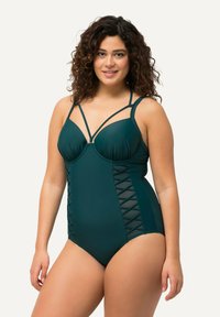 Maillot de bain une pièce teal avec bonnets structurés, bretelles réglables et détails croisées sur les côtés. Tissu lisse avec un léger éclat.