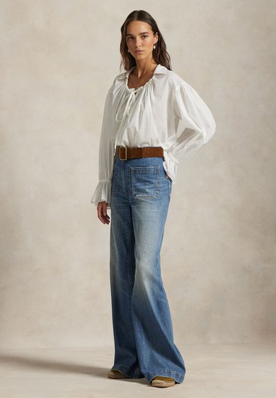 Femme portant un chemisier blanc à manches longues avec un col noué, un jean bleu délavé à jambes larges, une ceinture marron et des boucles d'oreilles, debout avec une main sur la hanche.