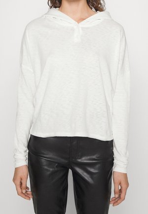 Kobieta w luźnej białej bluzie z długimi rękawami i czarnych skórzanych spodniach, stojąca na jednolitym jasnym tle.