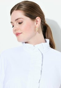 Top bianco con collo a imbuto e dettagli arricciati lungo la parte anteriore. Indossato con orecchini a cerchio dorati. Tessuto dalla texture liscia, sfondo illuminato.