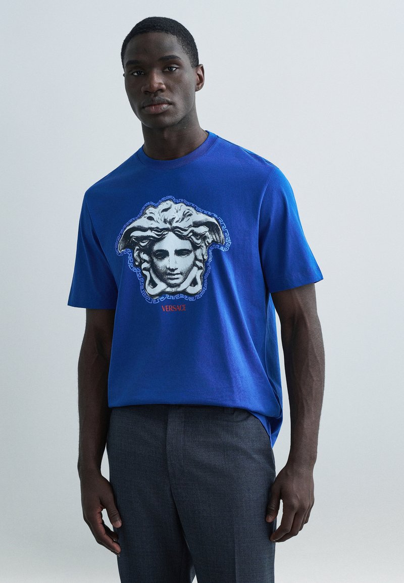 Modré bavlnené tričko s čiernobielou grafikou Medúzy a nápisom "Versace" pod ňou, kombinované so sivými prispôsobenými nohavicami.