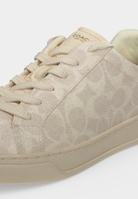 Μπεζ χαμηλό sneaker με υφασμάτινη υφή, ομοιόχρωμα κορδόνια και μπεζ σόλα, με διακριτικό μοτίβο λογότυπου Coach και ετικέτα στη γλώσσα με το λογότυπο.