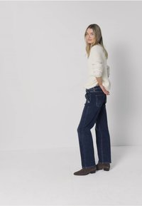 Suéter color crema claro, jeans azul oscuro de pierna ancha, botines marrones, de perfil, sobre un fondo neutro con estilo minimalista.