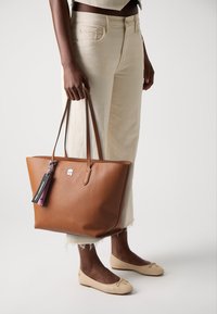 Sac tote en cuir marron à texture avec de longues poignées, arborant un logo argenté et une étiquette colorée. Porté avec un pantalon court de couleur claire et des ballerines.