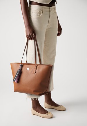 Borsa a spalla in pelle marrone con texture, dotata di manici lunghi, con un logo argento e un'etichetta colorata. Indossata con pantaloni cropped chiari e ballerine.