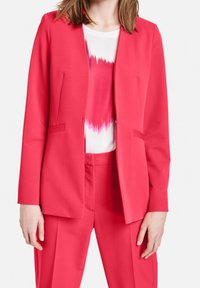 Mujer vistiendo un traje ajustado de un brillante color rosa, con un blazer y pantalones a juego, sobre una blusa blanca con un patrón abstracto en rosa.