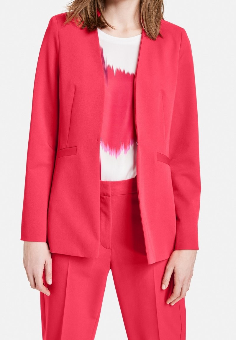 Mujer vistiendo un traje ajustado de un brillante color rosa, con un blazer y pantalones a juego, sobre una blusa blanca con un patrón abstracto en rosa.