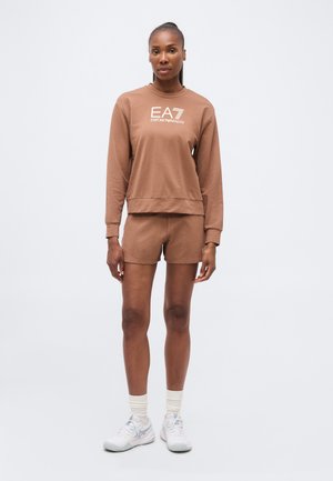 Femme debout portant un sweat-shirt marron EA7 Emporio Armani et un short assorti avec des chaussettes blanches et des baskets blanches sur un fond uni.