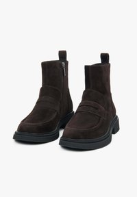 Tmavě hnědé semišové kotníkové boty s tlustými černými podrážkami, bočními zipy a poutky vzadu na bílém pozadí.