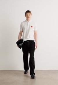 Hvid polo skjorte med en lille rød lap, sorte bukser og sorte sko. Modellen holder en sort clutch og står foran en ensfarvet baggrund.
