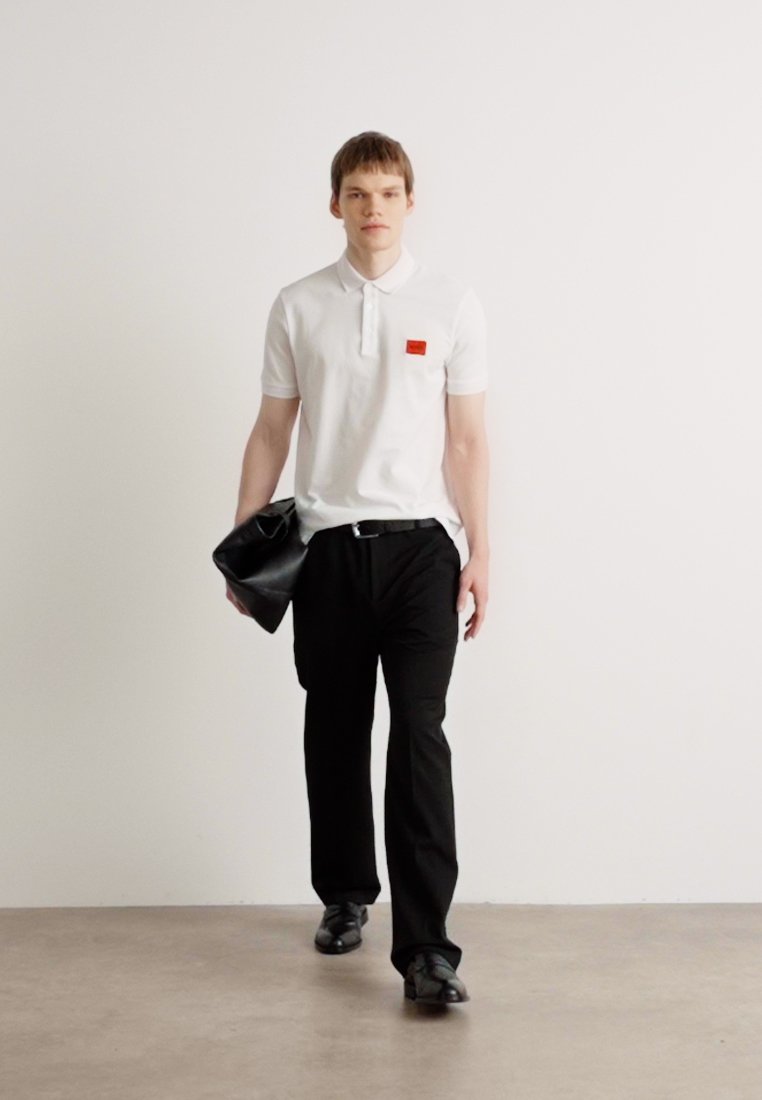Chemise polo blanche avec un petit patch rouge, pantalon noir et chaussures noires. Le mannequin tient une pochette noire, se tenant devant un fond uni.