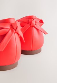 Paar rote Leder-Ballerinas mit Schleifen am Rücken und braunen flachen Sohlen auf hellem Hintergrund.