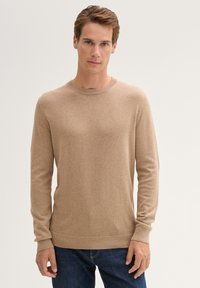 Beige gebreide sweater met een ronde hals, geribbelde manchetten en zoom. Model draagt het met donkerblauwe jeans, waardoor de textuur van de sweater benadrukt wordt.