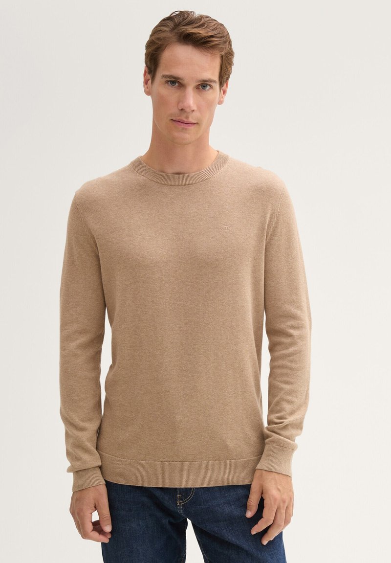 Beige gebreide sweater met een ronde hals, geribbelde manchetten en zoom. Model draagt het met donkerblauwe jeans, waardoor de textuur van de sweater benadrukt wordt.