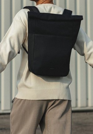 Personne portant un sac à dos noir à rabat roulé avec des bretelles rembourrées, un pull crème et un pantalon en velours côtelé beige, debout à l'extérieur contre un mur en métal.