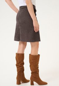 Kvinde iført en brun knælang nederdel og brune suede støvler med hæl til midt på læggen, stående på en hvid baggrund.