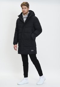 Sort pufferjakke med hætte, der har en lommeklap med lynlås, quiltet tekstur og et logo-patch nederst til venstre. Sammen med sorte bukser og hvide sneakers.
