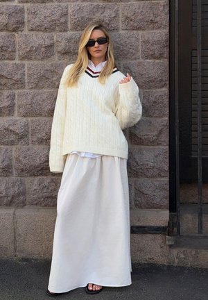 Dik wit kabelgebreide trui met een V-hals, gelaagd over een witte blouse en een vloeiende witte maxirok. Zwarte sandalen maken de look compleet.
