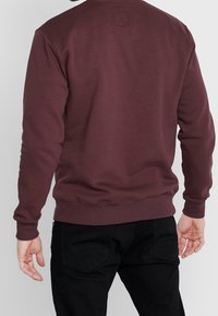 Sudadera burdeos con cuello redondo, mangas largas, puños y dobladillo acanalados. Diseño simple con un detalle de parche cuadrado en la parte de atrás.